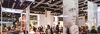 Besucher auf einer Messe, umgeben von modernem Standdesign und einem großen Porträt im Hintergrund.