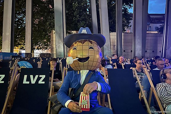 Leipziger Messe Maskottchen sitzt in einem Kinosaal im Freien mit Popcorn, Spaß und Unterhaltung für Besucher.