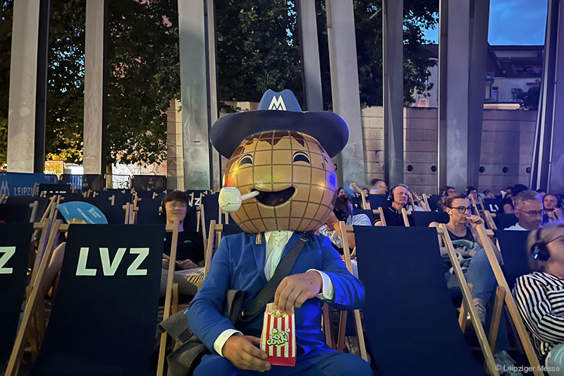 Leipziger Messe Maskottchen sitzt in einem Kinosaal im Freien mit Popcorn, Spaß und Unterhaltung für Besucher.